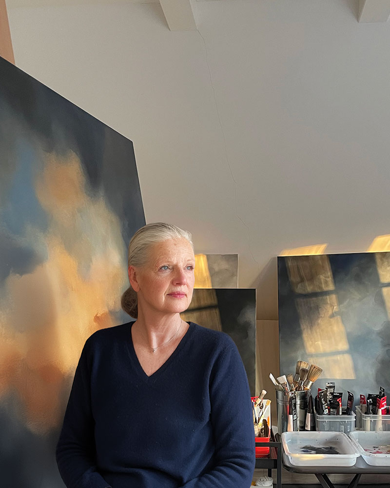 Die Künstlerin Christin von Wels sitzt in ihrem Kunstatelier vor einem großformatigen abstrakten Wolkengemälde. Das Bild zeigt atmosphärische Himmelslandschaften in Blau- und Goldtönen im warmen Sonnenlicht neben Pinsel und Farbtuben auf einem Arbeitstisch.