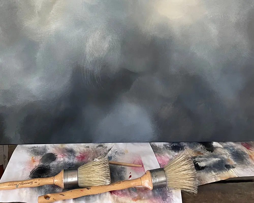 Zwei Rundpinsel liegen vor einem großformatigen Wolkenbild von Christin von Wels. Die Detailaufnahme zeigt die weichen Farbübergänge einer abstrakten Himmelslandschaft in Blau-, Grau- und Weißtönen sowie die Struktur der Acrylmalerei