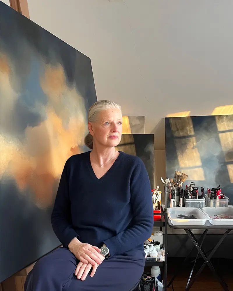 Künstlerin Christin von Wels sitzt in ihrem Kunstatelier vor einem großformatigen abstrakten Wolkengemälde. Das Bild zeigt atmosphärische Himmelslandschaften in Blau- und Goldtönen im warmen Sonnenlicht neben Pinsel und Farbtuben auf einem Arbeitstisch.
