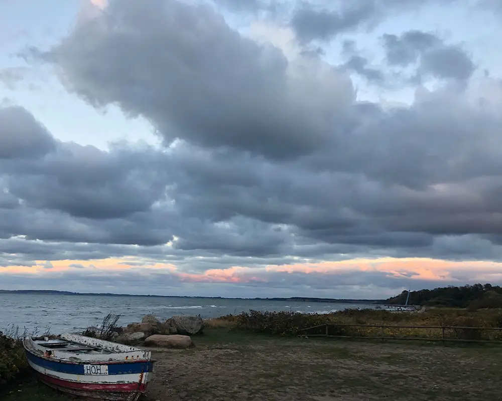 Atmosphärische Küstenszene in Hohwacht an der Ostsee: Ein altes Ruderboot am Ufer unter einem dramatischen Wolkenhimmel mit rosa Leuchten am Horizont – Inspiration für abstrakte Himmelslandschaften und Wolkenbilder der Künstlerin Christin von Wels